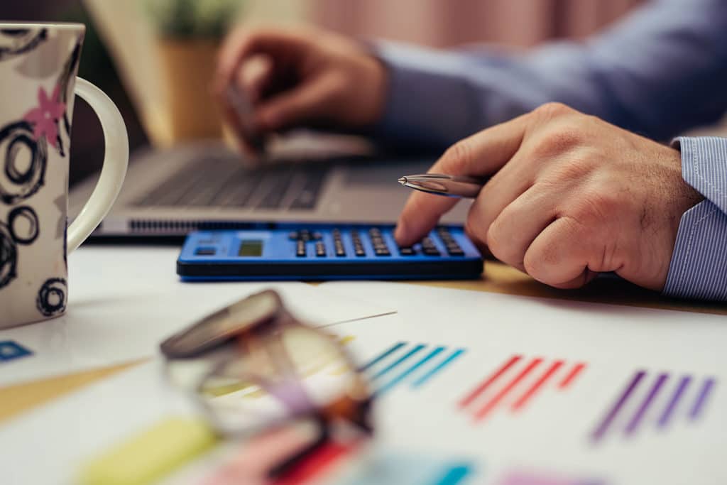 Male hands with one holding a pen while typing on a calculator, while the other is on the keyboard of a laptop