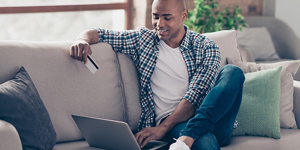 man sitting on couch smiling while working on his laptop and holding his credit card