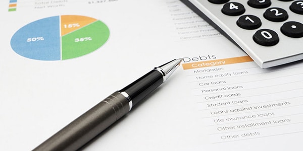 close-up of a pen on a desk with a calculator on top of a paper reading "Debts" with a pie chart near it